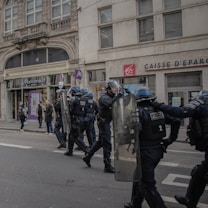A group of police officers in riot gear are moving in formation on a city street. They are equipped with helmets and shields, and appear to be in the middle of a coordinated action. The backdrop includes a building with large windows and visible shop names such as Marionnaud Paris and Caisse d'Epargne. Pedestrians are standing nearby, observing the scene.