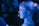 A person with their hair tied up is laughing in a dimly lit environment with a bluish hue. The image captures a candid moment, with other blurred figures in the background.