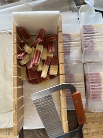 A collection of rectangular, ridged soap bars in shades of brown and cream are arranged on a paper towel within a wooden mold. A metal cutter with a wooden handle is placed on the edge of the mold. Additional wrapped soap bars are visible in the background.
