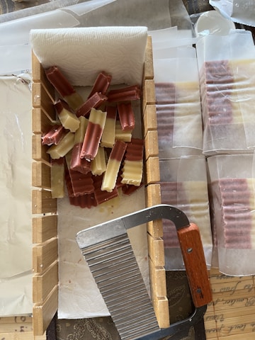 A collection of rectangular, ridged soap bars in shades of brown and cream are arranged on a paper towel within a wooden mold. A metal cutter with a wooden handle is placed on the edge of the mold. Additional wrapped soap bars are visible in the background.