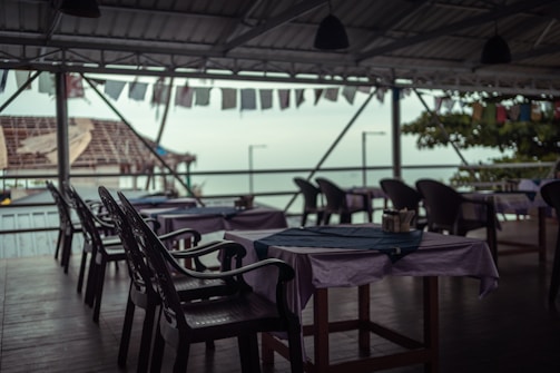 An open-air restaurant with empty tables and chairs, situated by the seaside. The tables are covered with cloths and the chairs are plastic. Overhead, the ceiling is metal and decorated with strings of flags. The background shows a partly visible ocean with some greenery and a structure resembling a roof beyond the railing.