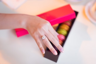 A hand with a ring displaying a diamond placed delicately on the ring finger. The fingernails are manicured, and the hand is positioned just above an open box of colorful macarons.