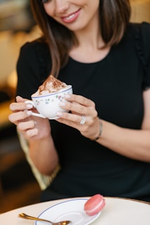 A woman with a warm smile holds a delicate floral cup filled with a frothy beverage topped with cocoa powder. She is wearing a black top and has brown hair. A pink macaron sits on a white saucer with a blue rim, accompanied by a spoon on a light-colored table.