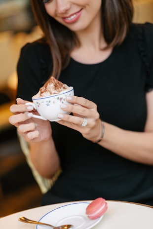 A woman with a warm smile holds a delicate floral cup filled with a frothy beverage topped with cocoa powder. She is wearing a black top and has brown hair. A pink macaron sits on a white saucer with a blue rim, accompanied by a spoon on a light-colored table.