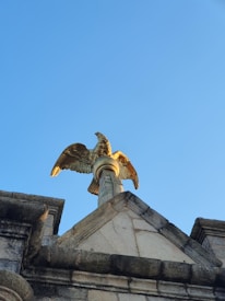 A stone building features a decorative golden eagle perched on top. The eagle is positioned on a pedestal at the peak of the structure, with its wings partially spread. The sky is clear and blue, providing a bright backdrop.
