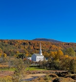 A white church with a tall steeple is nestled among a vibrant landscape of autumn foliage, composed of yellow, orange, and green hues. Behind the church, a forested hill rises, culminating in a distant mountain peak under a clear blue sky.