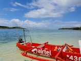 A small red boat with 'Airtel Money' branding is anchored on clear, shallow waters near a serene beach. The ocean stretches out to the horizon, with a backdrop of lush green trees on a distant island. The sky above is partly cloudy, adding depth to the tranquil setting.