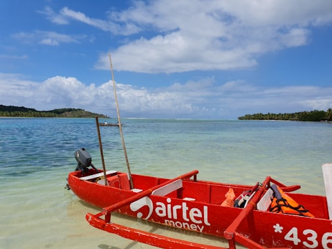 A small red boat with 'Airtel Money' branding is anchored on clear, shallow waters near a serene beach. The ocean stretches out to the horizon, with a backdrop of lush green trees on a distant island. The sky above is partly cloudy, adding depth to the tranquil setting.