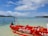 A small red boat with 'Airtel Money' branding is anchored on clear, shallow waters near a serene beach. The ocean stretches out to the horizon, with a backdrop of lush green trees on a distant island. The sky above is partly cloudy, adding depth to the tranquil setting.