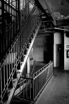 A dimly lit indoor space featuring a metal staircase with railings, casting shadows on the wall. The environment appears aged and neglected, with a cracked ceiling and flaking paint. A small doorway leads to another room, and there's graffiti on the wall with the words 'ela &eacute; um'.