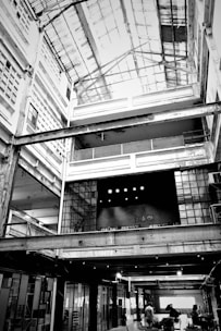 A large, industrial-style interior space with an open ceiling structure. The building features multiple levels with exposed beams and girders, and there is a significant use of glass and metal in the construction. The word 'Fabrica' is visible on a textured wall in the center, suggesting the space may be used for creative or manufacturing purposes. People are visible at the ground level, giving the space a sense of scale.