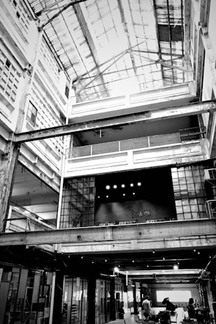 A large, industrial-style interior space with an open ceiling structure. The building features multiple levels with exposed beams and girders, and there is a significant use of glass and metal in the construction. The word 'Fabrica' is visible on a textured wall in the center, suggesting the space may be used for creative or manufacturing purposes. People are visible at the ground level, giving the space a sense of scale.