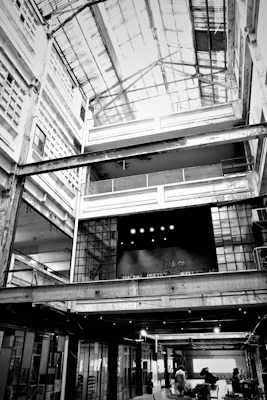 A large, industrial-style interior space with an open ceiling structure. The building features multiple levels with exposed beams and girders, and there is a significant use of glass and metal in the construction. The word 'Fabrica' is visible on a textured wall in the center, suggesting the space may be used for creative or manufacturing purposes. People are visible at the ground level, giving the space a sense of scale.