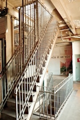 An industrial-style staircase with metal railings and a mesh enclosure is visible, leading to an upper floor in a building. The surrounding area appears slightly worn, with exposed pipes and cracked paint. A phrase in Portuguese, 'ela é um elo,' is written on a nearby wall, adding an artistic touch. Some greenery can be seen in the background.