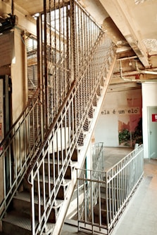 An industrial-style staircase with metal railings and a mesh enclosure is visible, leading to an upper floor in a building. The surrounding area appears slightly worn, with exposed pipes and cracked paint. A phrase in Portuguese, 'ela &eacute; um elo,' is written on a nearby wall, adding an artistic touch. Some greenery can be seen in the background.