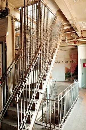 An industrial-style staircase with metal railings and a mesh enclosure is visible, leading to an upper floor in a building. The surrounding area appears slightly worn, with exposed pipes and cracked paint. A phrase in Portuguese, 'ela &eacute; um elo,' is written on a nearby wall, adding an artistic touch. Some greenery can be seen in the background.
