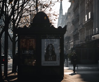 A street scene with a kiosk displaying advertisements in front of a row of Parisian-style buildings. The background features a tall, pointed church spire partially obscured by mist. Tree branches with sparse autumn leaves frame the scene, and a few pedestrians are visible, casting long shadows on the pavement.