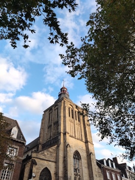 A stone church tower stands prominently against a backdrop of a bright blue sky with scattered clouds. The architecture features Gothic-style elements, including tall, narrow windows with a pointed arch design. Surrounding the tower are the tops of trees, with leafy branches framing the view. Adjacent buildings have sloped rooftops and brick facades, enhancing the historical ambiance.
