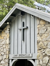 A rustic stone structure features a wooden cross mounted on the front, above a sign reading 'Camp Greenville'. The building is made of light gray wood and rough stone, with a pointed roof partially covered by nearby tree branches.