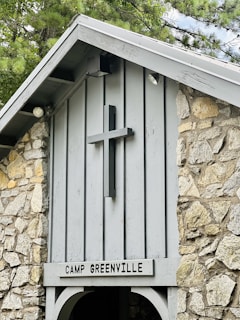 A rustic stone structure features a wooden cross mounted on the front, above a sign reading 'Camp Greenville'. The building is made of light gray wood and rough stone, with a pointed roof partially covered by nearby tree branches.