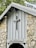 A rustic stone structure features a wooden cross mounted on the front, above a sign reading 'Camp Greenville'. The building is made of light gray wood and rough stone, with a pointed roof partially covered by nearby tree branches.