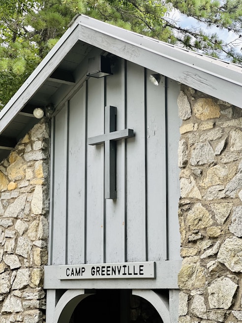 A rustic stone structure features a wooden cross mounted on the front, above a sign reading 'Camp Greenville'. The building is made of light gray wood and rough stone, with a pointed roof partially covered by nearby tree branches.