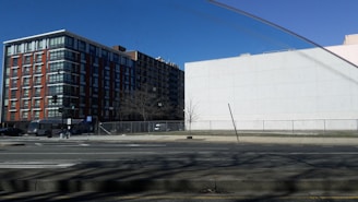 A city street scene with a tall modern apartment building on the left, characterized by glass windows and red brick facades. To the right, a large blank white wall stands adjacent to an empty lot enclosed by a wire fence. A bare tree and a slightly leaning street sign are visible on the sidewalk. The sky is clear and blue, indicating a sunny day.