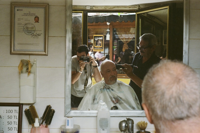 A man is having his hair cut in a barbershop. He is seated in front of a mirror, with a cape draped around his shoulders. A barber is working, holding a clipper. The reflection shows another person taking a photograph, holding a camera. The barbershop has a certificate or license on the wall and several grooming tools like brushes are visible.