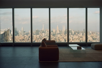 A spacious, modern living room with a large window overlooking an expansive cityscape. The furniture includes a sofa and a glass coffee table placed on a minimalist rug, highlighting the sleek design of the interior. The view showcases numerous skyscrapers under a partly cloudy sky, creating an atmosphere of urban sophistication.