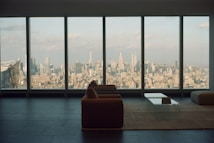 A spacious, modern living room with a large window overlooking an expansive cityscape. The furniture includes a sofa and a glass coffee table placed on a minimalist rug, highlighting the sleek design of the interior. The view showcases numerous skyscrapers under a partly cloudy sky, creating an atmosphere of urban sophistication.