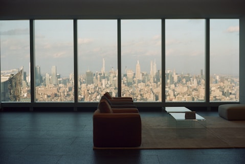 A spacious, modern living room with a large window overlooking an expansive cityscape. The furniture includes a sofa and a glass coffee table placed on a minimalist rug, highlighting the sleek design of the interior. The view showcases numerous skyscrapers under a partly cloudy sky, creating an atmosphere of urban sophistication.