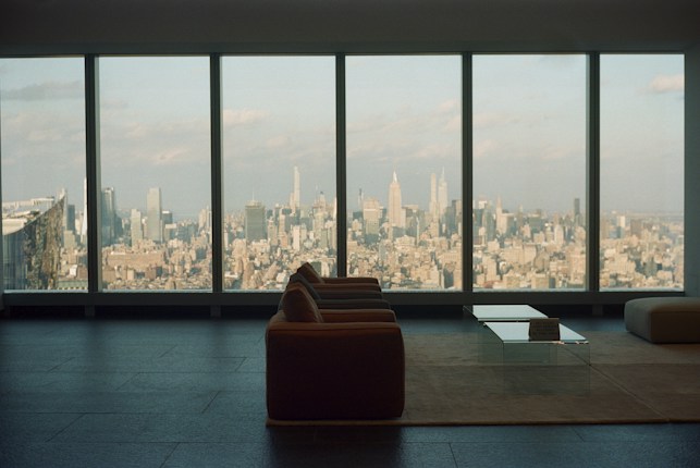 A spacious, modern living room with a large window overlooking an expansive cityscape. The furniture includes a sofa and a glass coffee table placed on a minimalist rug, highlighting the sleek design of the interior. The view showcases numerous skyscrapers under a partly cloudy sky, creating an atmosphere of urban sophistication.