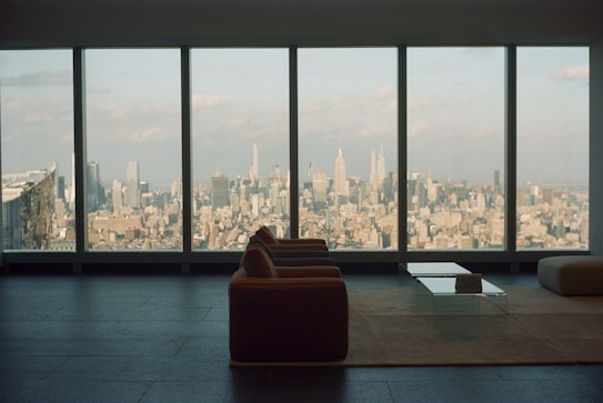 A spacious, modern living room with a large window overlooking an expansive cityscape. The furniture includes a sofa and a glass coffee table placed on a minimalist rug, highlighting the sleek design of the interior. The view showcases numerous skyscrapers under a partly cloudy sky, creating an atmosphere of urban sophistication.