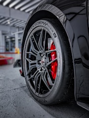 A close-up of a car wheel with an intricate black alloy rim design and visible brand markings on the tire. The red brake caliper provides a striking contrast against the black wheel. The car is parked on a concrete surface with structural elements visible in the background.