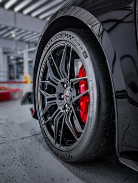 A close-up of a car wheel with an intricate black alloy rim design and visible brand markings on the tire. The red brake caliper provides a striking contrast against the black wheel. The car is parked on a concrete surface with structural elements visible in the background.