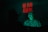 A man with clown makeup is illuminated by a green light, wearing a polka dot shirt. In the background, there is a neon sign with red lettering that says 'WE RUN ATHENS'. Another person, partially visible, appears to be sitting opposite the clown.