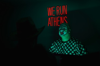 A man with clown makeup is illuminated by a green light, wearing a polka dot shirt. In the background, there is a neon sign with red lettering that says 'WE RUN ATHENS'. Another person, partially visible, appears to be sitting opposite the clown.