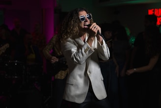 A person with long hair wearing a white jacket and sunglasses is passionately singing into a microphone. The dimly lit room appears to be a stage or a club setting with colorful lights creating an atmospheric vibe. A crowd is visible in the background, hinting at a lively performance.