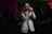 A person with long hair wearing a white jacket and sunglasses is passionately singing into a microphone. The dimly lit room appears to be a stage or a club setting with colorful lights creating an atmospheric vibe. A crowd is visible in the background, hinting at a lively performance.