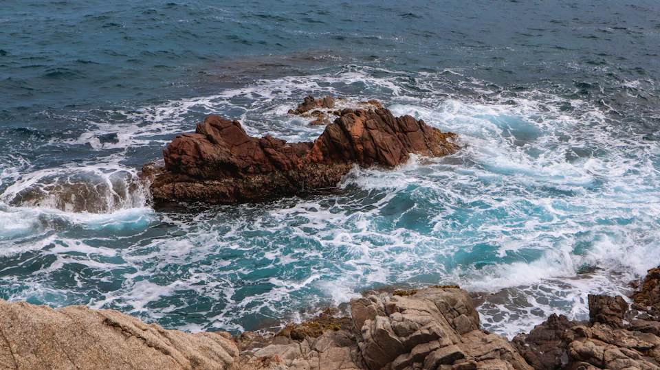 The image depicts a rocky coastline with a large, jagged rock formation on the left side and smaller rocks scattered across the right side. The water surrounding the rocks is a deep blue color, indicating that it's likely during the day when the sun is shining brightly. The sky above mirrors the ocean below in its light blue hue.