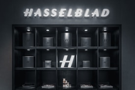 A display shelf with multiple compartments holds several black boxes, each featuring the letter 'H' on them. Above the shelf, the name 'Hasselblad' is illuminated. The boxes are evenly spaced, and there is also a prominent glowing 'H' in one of the compartments. The lighting is predominantly dark, with spotlights highlighting the boxes.