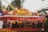 A vibrant outdoor food stall named Broer's Currywurst is bustling with people. The stall is decorated in bright red and features large signage with images of sausages and fries. It is set in a lively area, with amusement park rides visible in the background.