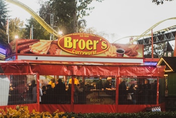A vibrant outdoor food stall named Broer's Currywurst is bustling with people. The stall is decorated in bright red and features large signage with images of sausages and fries. It is set in a lively area, with amusement park rides visible in the background.