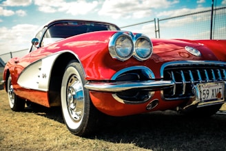 A classic red sports car, likely a vintage Corvette, is parked on a grassy area. The car features a gleaming chrome front grille, dual headlights, and a California license plate. The image is taken from a low angle, emphasizing the car's sleek design and shiny surface. In the background, a chain-link fence and a partly cloudy sky can be seen.