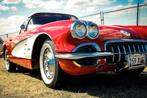 A classic red sports car, likely a vintage Corvette, is parked on a grassy area. The car features a gleaming chrome front grille, dual headlights, and a California license plate. The image is taken from a low angle, emphasizing the car's sleek design and shiny surface. In the background, a chain-link fence and a partly cloudy sky can be seen.