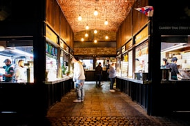 An indoor market corridor with a high brick ceiling lit by hanging bulbs. Food stalls line each side, with people preparing and serving dishes. The floor is made of cobblestone, adding a rustic feel to the vibrant environment. There are plants and menu boards above the counters, contributing to a lively atmosphere.