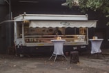 A food truck with a canopy is parked against a dark building. It is decorated with images of fish and sandwiches, suggesting it serves seafood. Two tall tables covered with white cloth are placed in front of the truck, each holding napkins and condiments. The area is littered with a few fallen leaves, and there is a tree partially visible to the right.