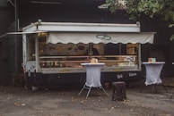 A food truck with a canopy is parked against a dark building. It is decorated with images of fish and sandwiches, suggesting it serves seafood. Two tall tables covered with white cloth are placed in front of the truck, each holding napkins and condiments. The area is littered with a few fallen leaves, and there is a tree partially visible to the right.