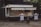 A food truck with a canopy is parked against a dark building. It is decorated with images of fish and sandwiches, suggesting it serves seafood. Two tall tables covered with white cloth are placed in front of the truck, each holding napkins and condiments. The area is littered with a few fallen leaves, and there is a tree partially visible to the right.
