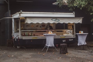 A food truck with a canopy is parked against a dark building. It is decorated with images of fish and sandwiches, suggesting it serves seafood. Two tall tables covered with white cloth are placed in front of the truck, each holding napkins and condiments. The area is littered with a few fallen leaves, and there is a tree partially visible to the right.